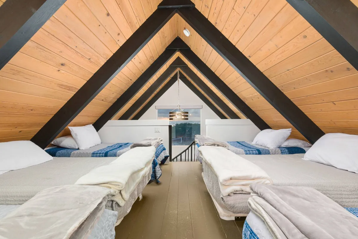 A tranquil sleeping area beneath a sloped wooden ceiling, with multiple beds dressed in soft linens and a large window that lets in natural light. The mood is serene, inviting, and ideal for relaxation or daydreaming.