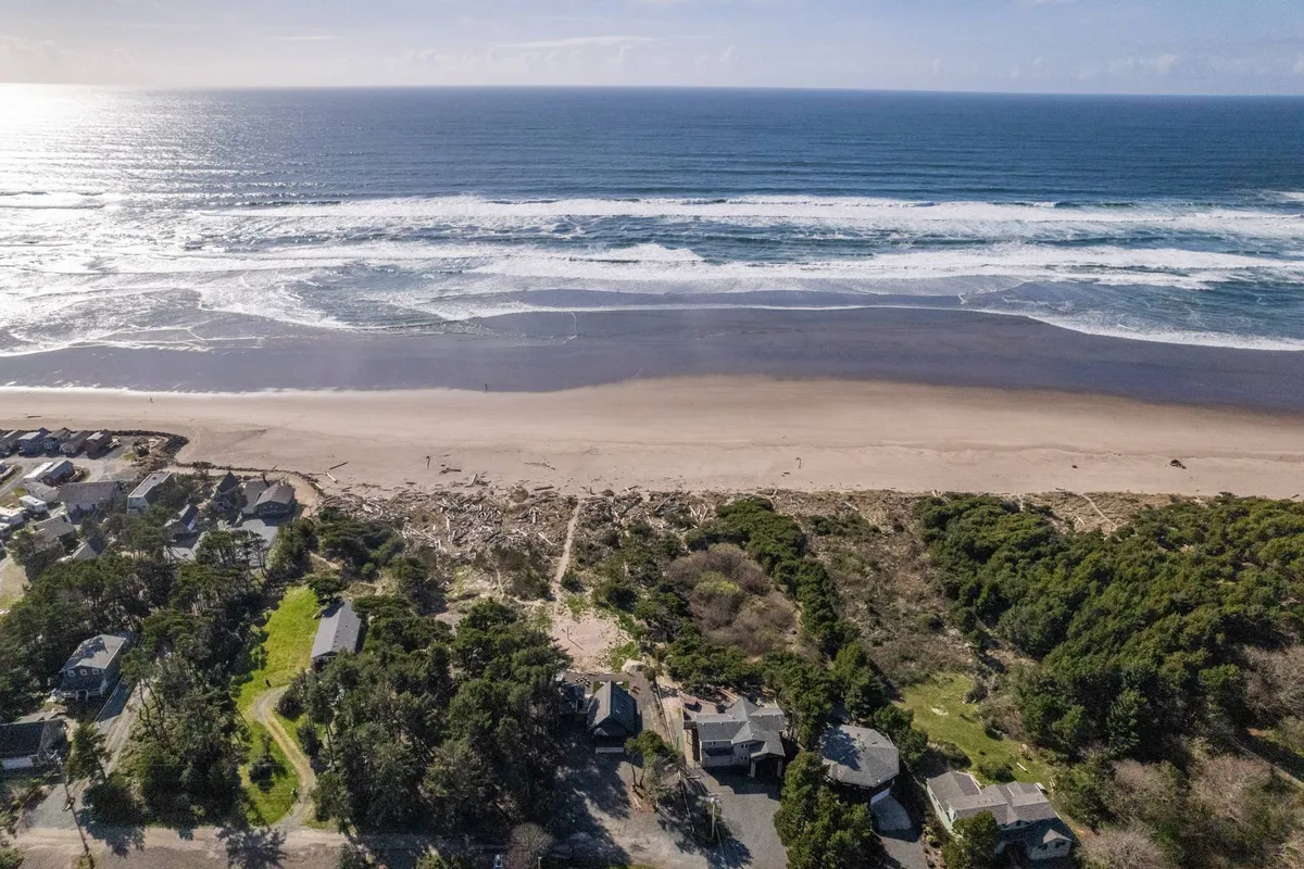 A sweeping view of the beach and ocean waves, framed by coastal forest and distant houses, capturing direct beach access and the natural beauty steps away from the property.