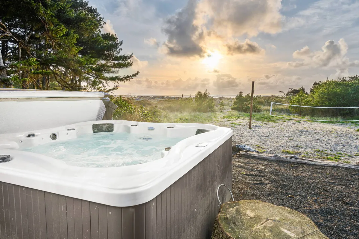 The hero image showcases the exterior of a charming A-frame house nestled among coastal trees, with a private hot tub overlooking the beach. The sun is setting, casting warm light across the sand, and guests can be seen relaxing in the hot tub, playing volleyball, and strolling along the shore, all surrounded by the peaceful beauty of nature.