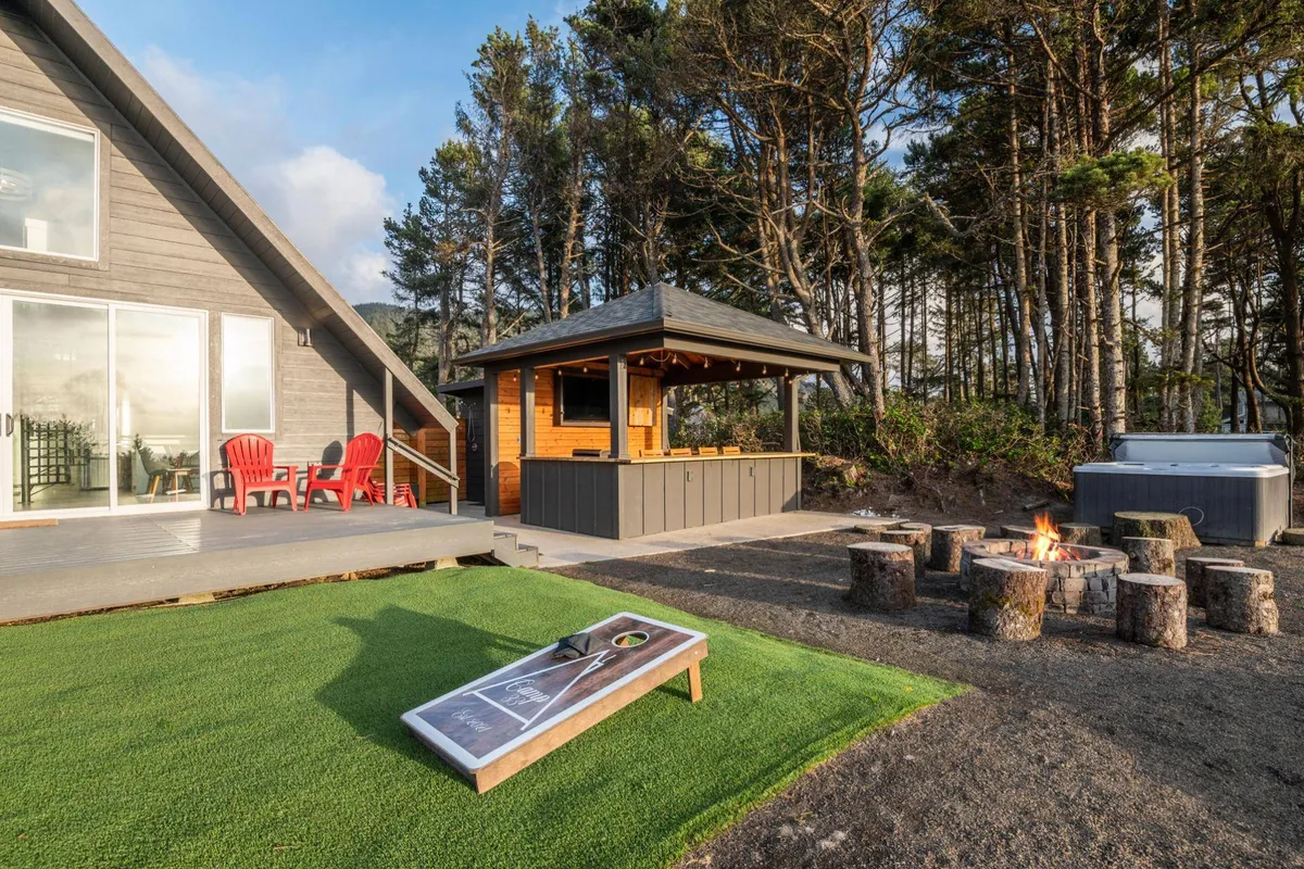 A vibrant exterior shot featuring red Adirondack chairs around a fire pit, a visible outdoor bar, and cornhole game boards set up on the lawn. The arrangement encourages socializing, playing games, and relaxing outdoors as a group.