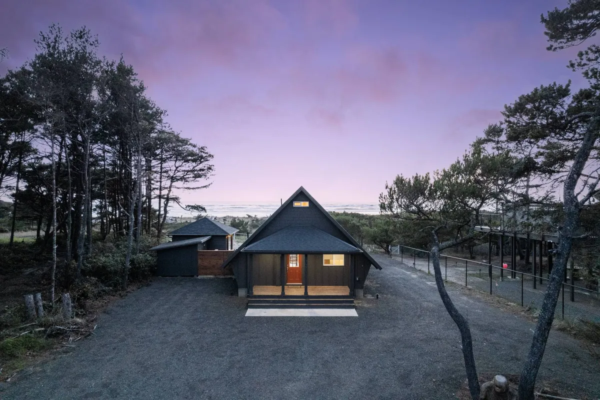 The A-frame cabin's wooden entrance is nestled among towering trees, with a welcoming porch perfect for enjoying nature. The tranquil vibe and natural setting offer a sense of privacy and connection to the outdoors.
