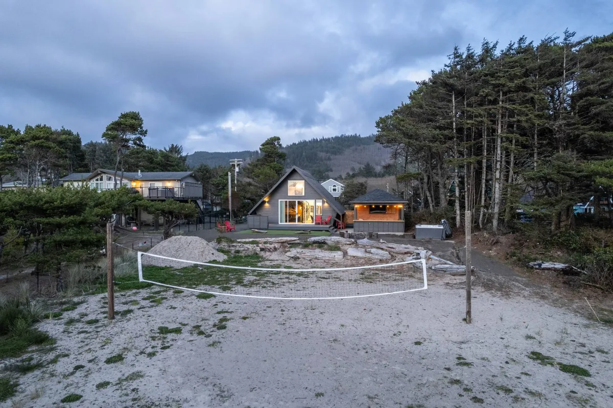 A casual, inviting view of an A-frame house with a sand volleyball court in the foreground and an outdoor seating area for groups to gather. The scene highlights the court set up for play, a fire pit for group gatherings, and ample space for outdoor meals.