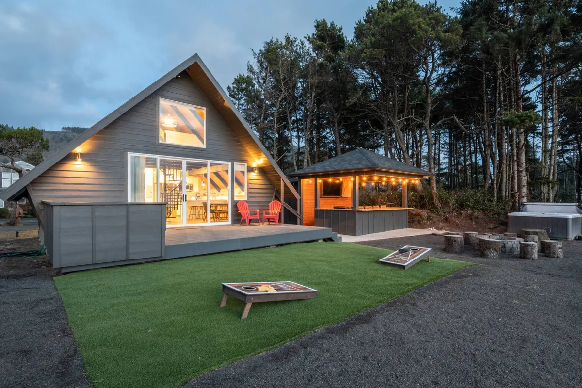 This image shows the A-frame house with a welcoming outdoor seating area, a sprawling grassy lawn, and a covered barbecue area. The wide open space is bordered by trees and bathed in warm sunlight, signaling a pet-friendly environment with plenty of space for pets to play safely. The setup is ideal for relaxing, barbecuing, and gathering outdoors.