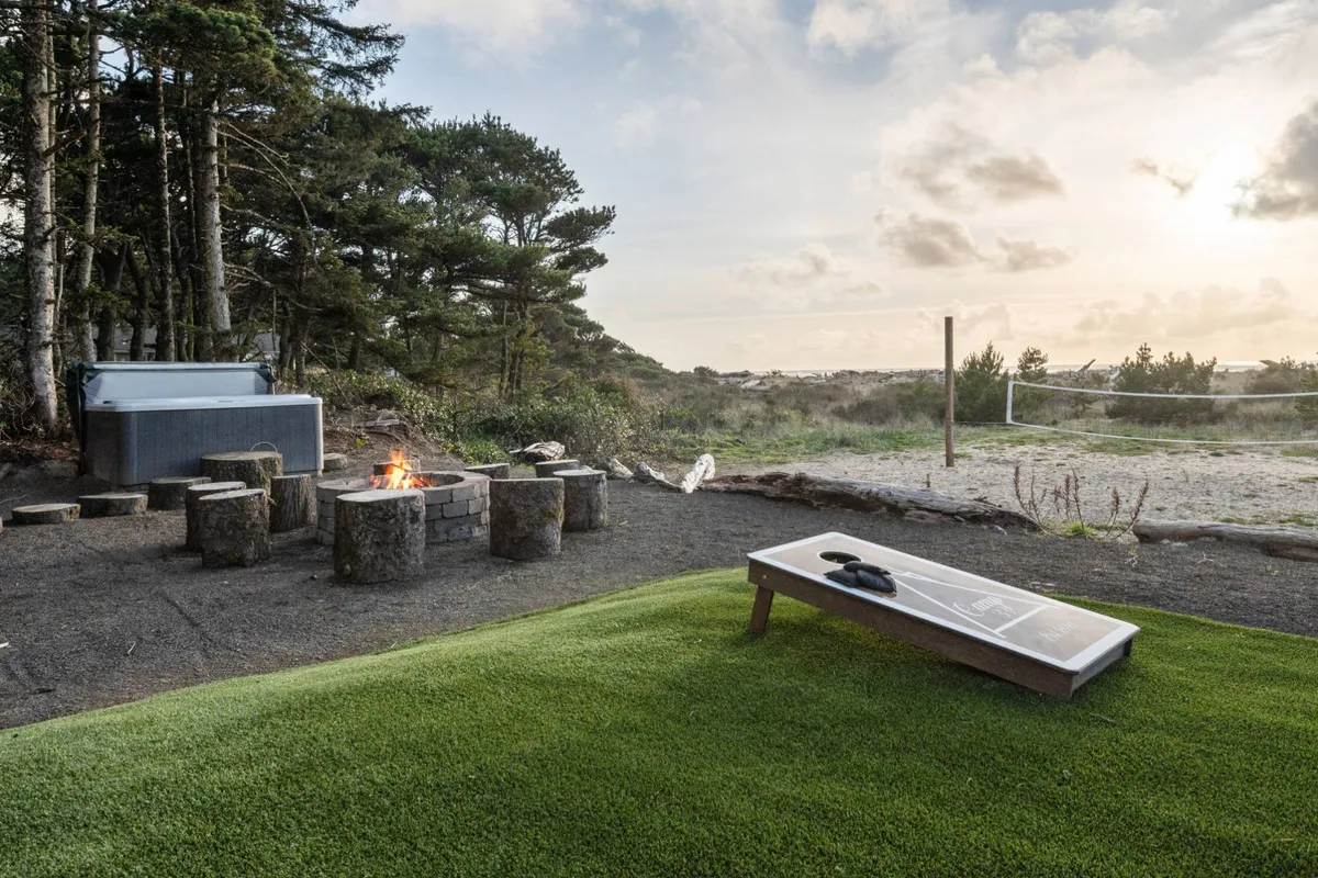 This exterior shot captures a vibrant backyard scene featuring a fire pit ringed by cozy seating, a private hot tub off to the side, a well-kept lawn, and a volleyball net ready for action—all under the warm glow of sunset.