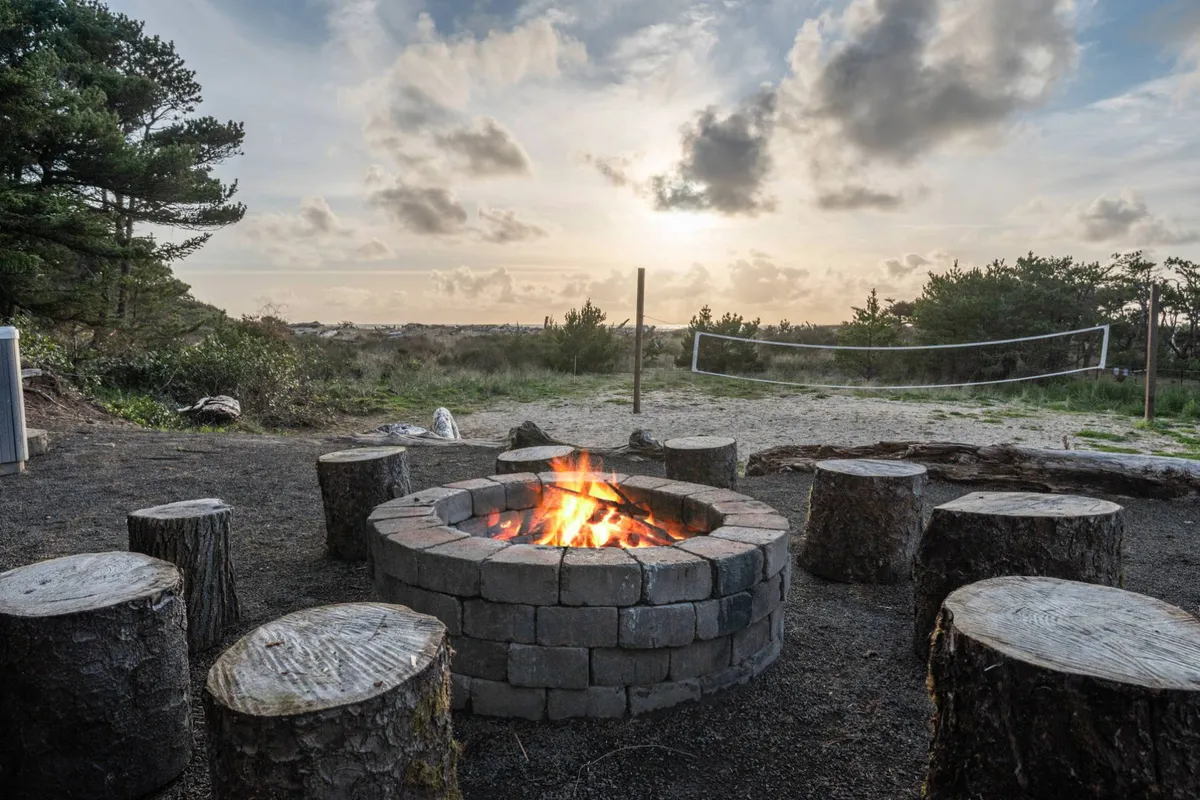 A cozy fire pit area with wooden seating and a volleyball net nearby, all set against a stunning sunset backdrop. The space feels inviting for group gatherings, games, and relaxation as the day ends.