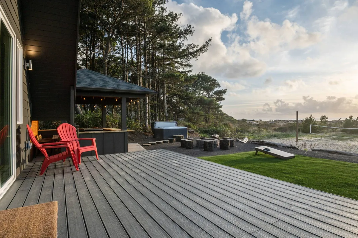 A tranquil outdoor scene with a covered gazebo, red chairs, a wooden deck, outdoor kitchen area, fire pit, and a green lawn. The space is set up for outdoor dining, relaxation, and group gatherings, with the Pacific Ocean just beyond.
