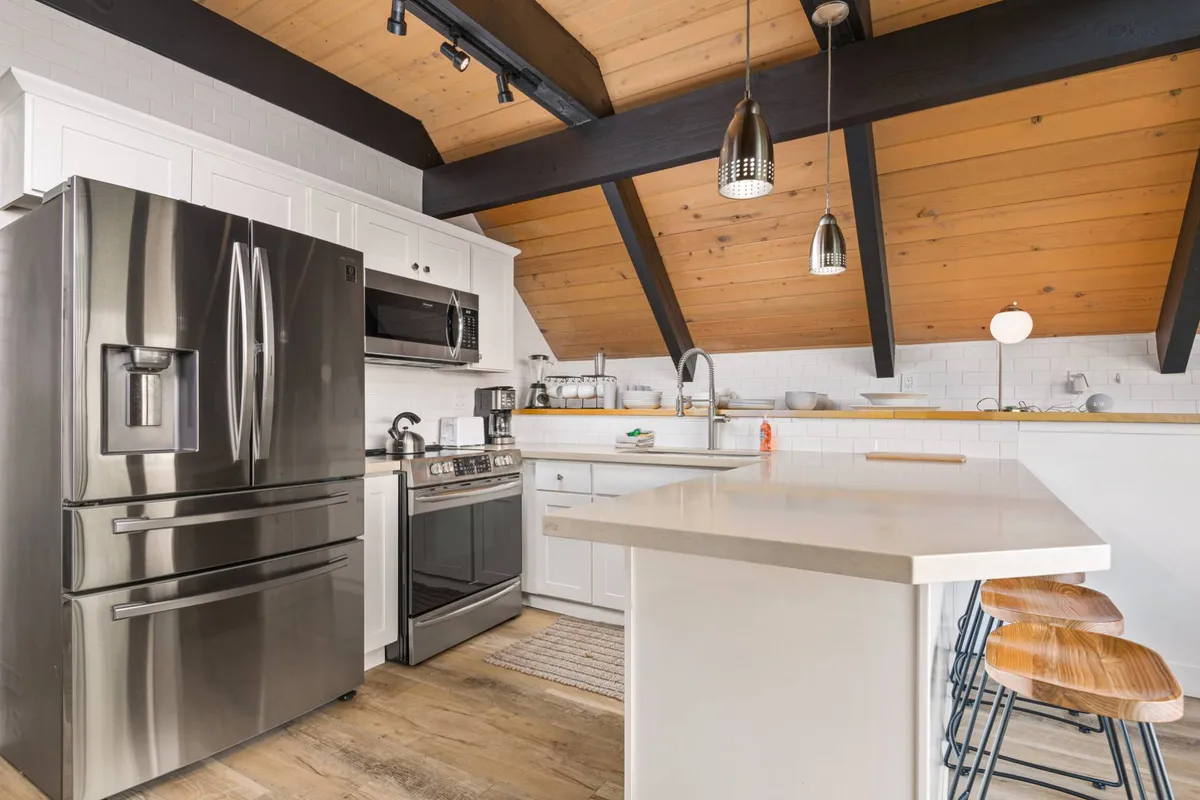 A spacious, modern kitchen with stainless steel appliances, a large kitchen island serving as a breakfast bar, and exposed wooden beams overhead. The area is bright and welcoming, inviting both cooking and social gatherings.