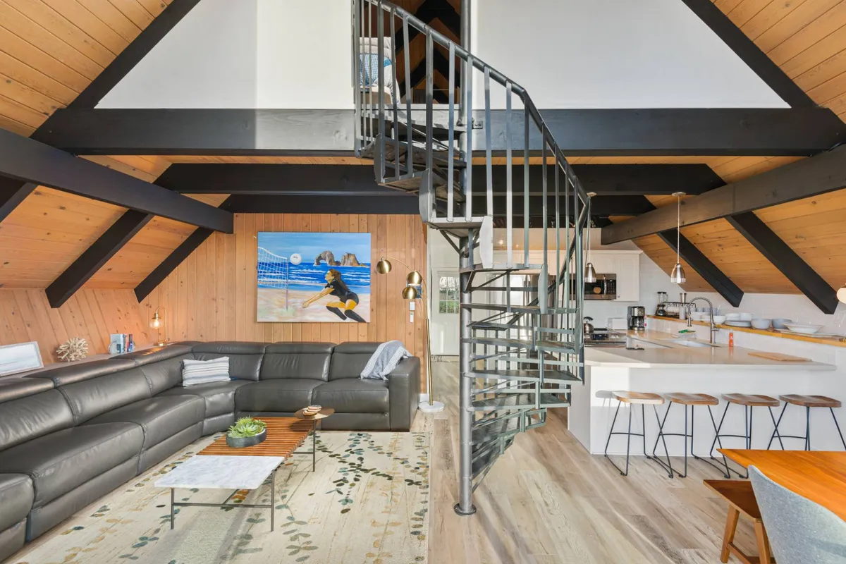 This image captures the open-concept living area, featuring a large, cozy sectional sofa as the social hub of the room. The eye-catching spiral staircase stands out, hinting at the loft area above, while natural light brightens the space. Artwork and a kitchen island in the background add depth and vibrancy, supporting group activities and togetherness.