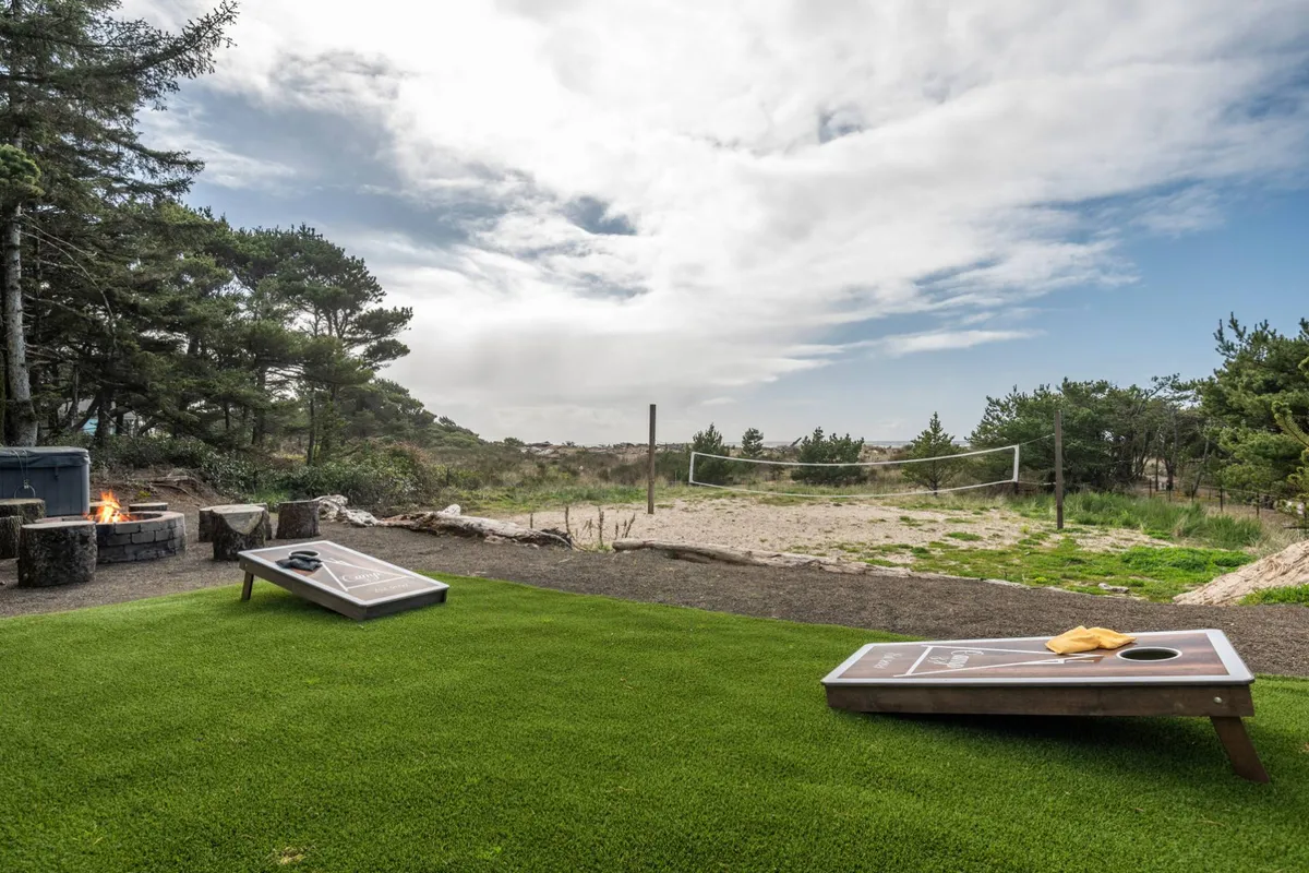 A playful and communal backyard scene with cornhole boards, a fire pit surrounded by chairs, a volleyball net, and lush greenery, suggesting opportunities for games, gatherings, and relaxation.