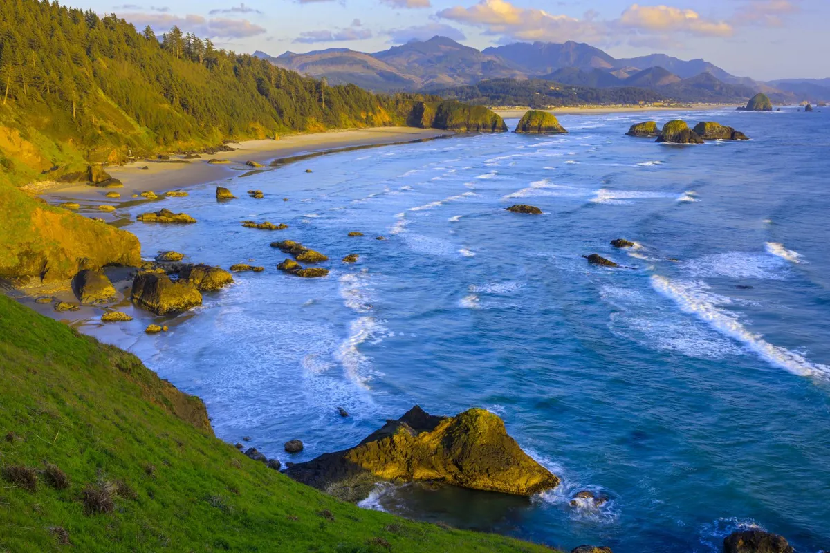 A sweeping view of a pristine beach with gentle ocean waves, dramatic rock formations, and lush mountain and forest backdrops. The setting sun casts a warm glow over the water, creating an inspiring and tranquil atmosphere.