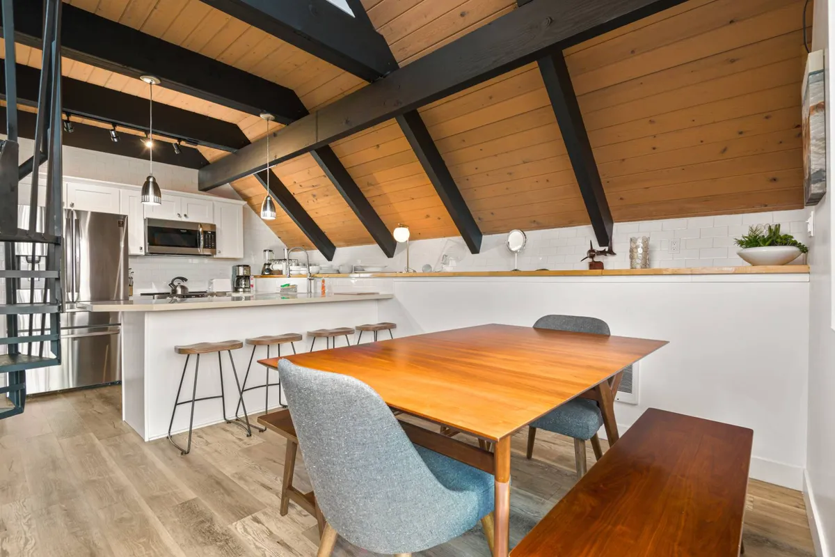 A warm, light-filled dining space featuring a bench-style dining table, a spiral staircase, and a kitchen island in the background. The wooden beams and inviting layout create an atmosphere ideal for group meals, board games, and laughter.