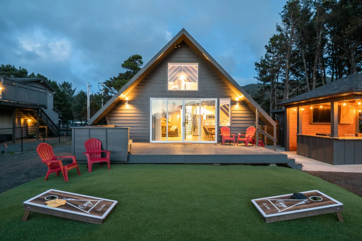 An exterior view showcasing the A-frame house, bright red chairs, outdoor game boards, and a landscaped green lawn. The setup encourages outdoor play, relaxation, and social activities, all in a welcoming and pet-appropriate setting.